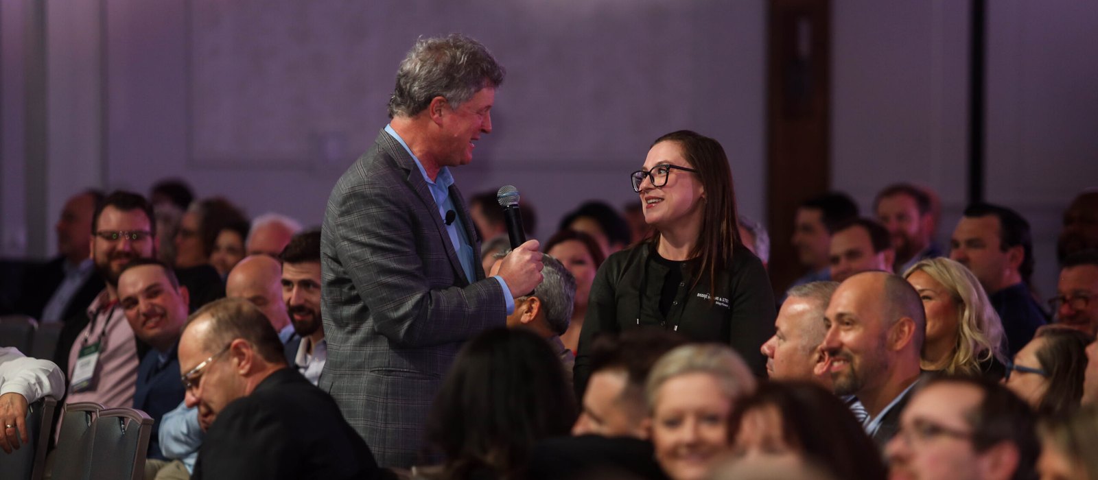 Conference emcee Greg Schwem holding a microphone for a laughing attendee during a corporate general session